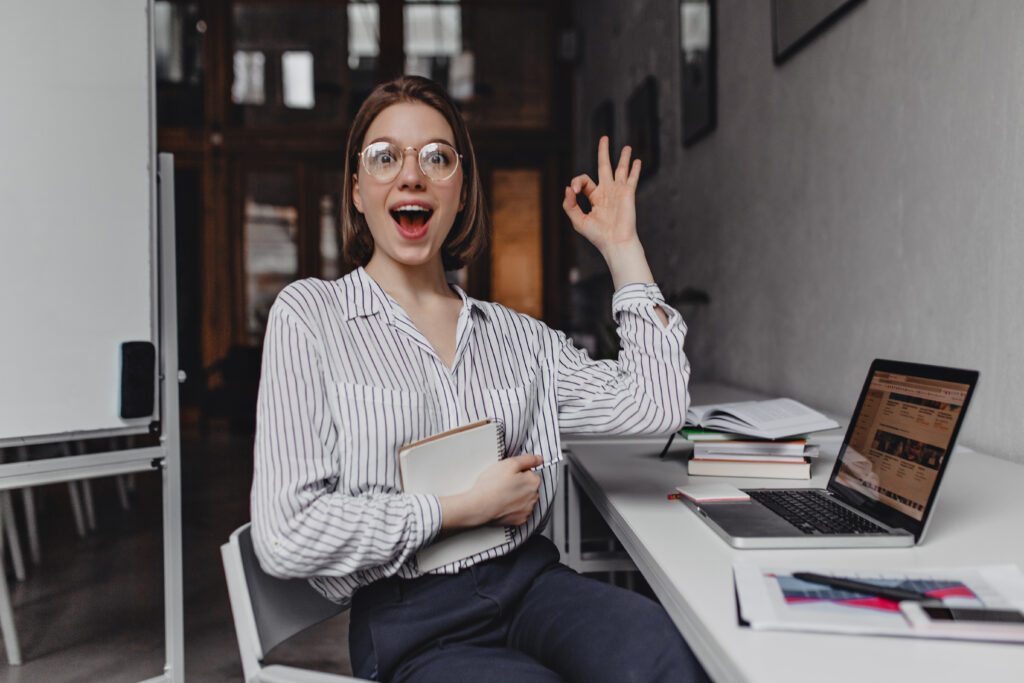 Joyful girl office worker shows OK sign. Portrait of woman in pants and light blouse at workplace.